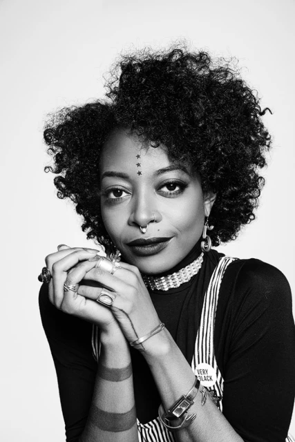 Black and white portrait of a woman with curly hair, wearing jewelry and a striped outfit, posing against a plain white backdrop.