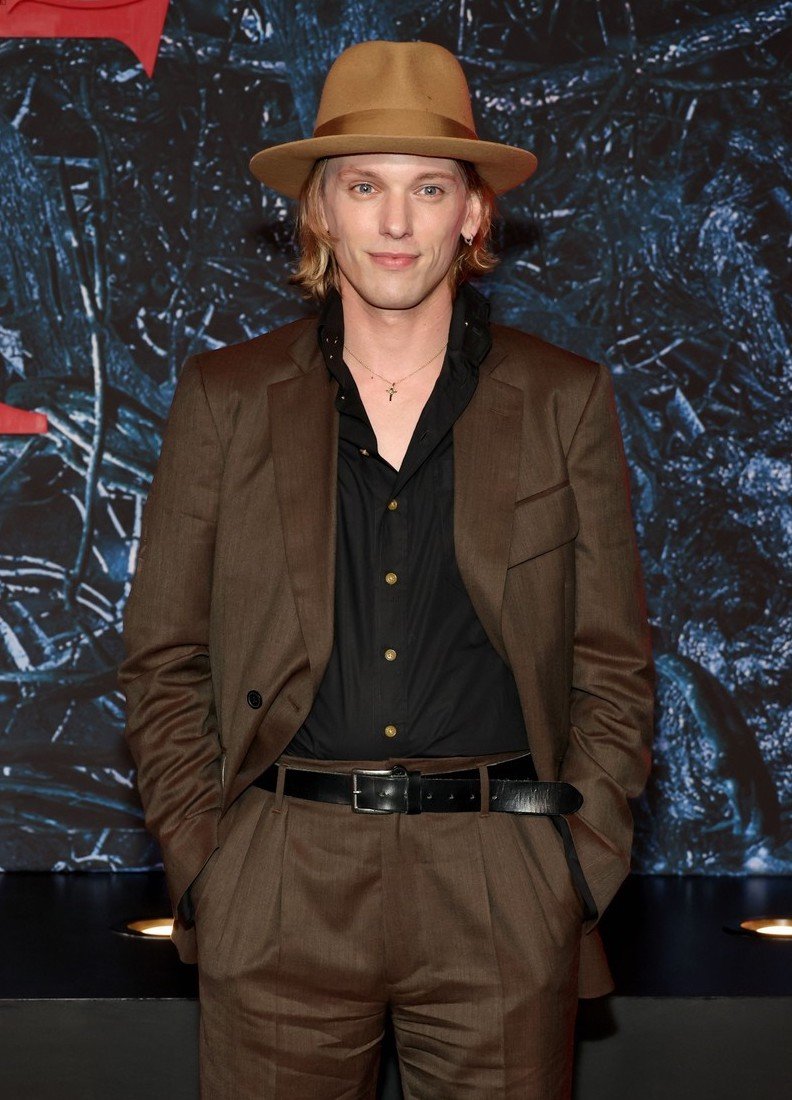 Man wearing a brown suit, black shirt, and fedora hat posing against a dark textured backdrop in New York.