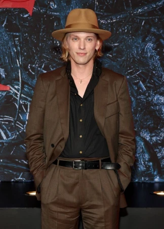 Man wearing a brown suit, black shirt, and fedora hat posing against a dark textured backdrop in New York.