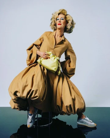 Older woman with voluminous blonde hair wearing a camel balloon dress and holding a pale yellow handbag against a white background