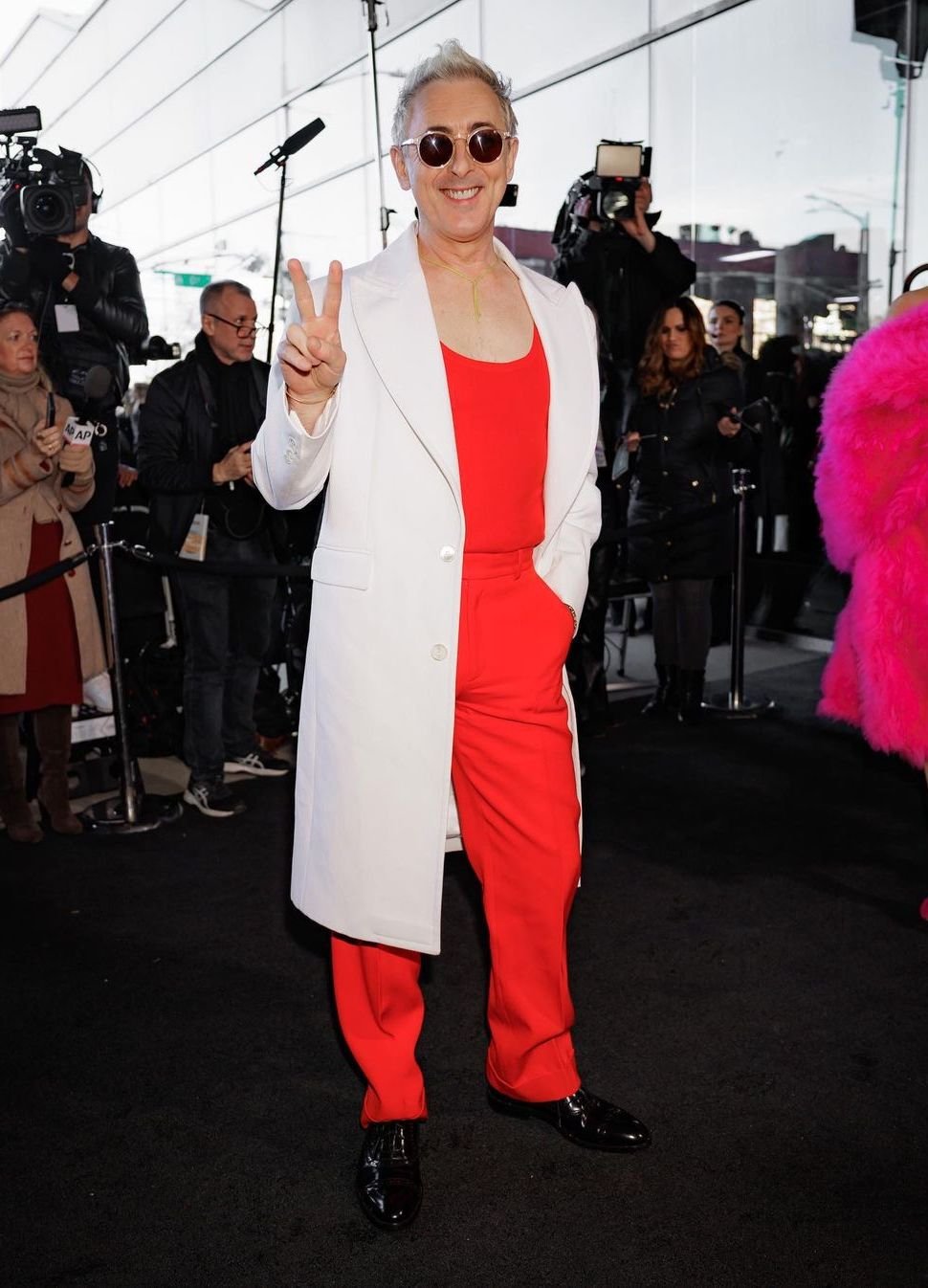 Alan Cumming poses on a black carpet in a red jumpsuit and white coat, flashing a peace sign with photographers and onlookers in background.