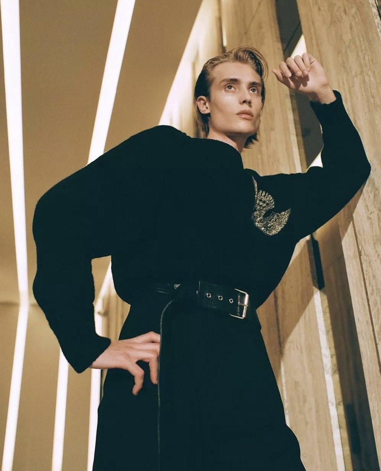 Low-angle shot of a male model in a black sweater with a bird brooch leaning against a wooden paneled wall.