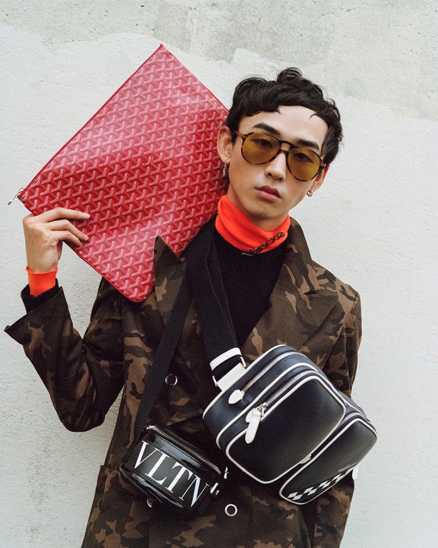 A stylish individual poses outdoors in a camo jacket, accessorized with a Goyard clutch and Valentino crossbody bags, set against a neutral wall.