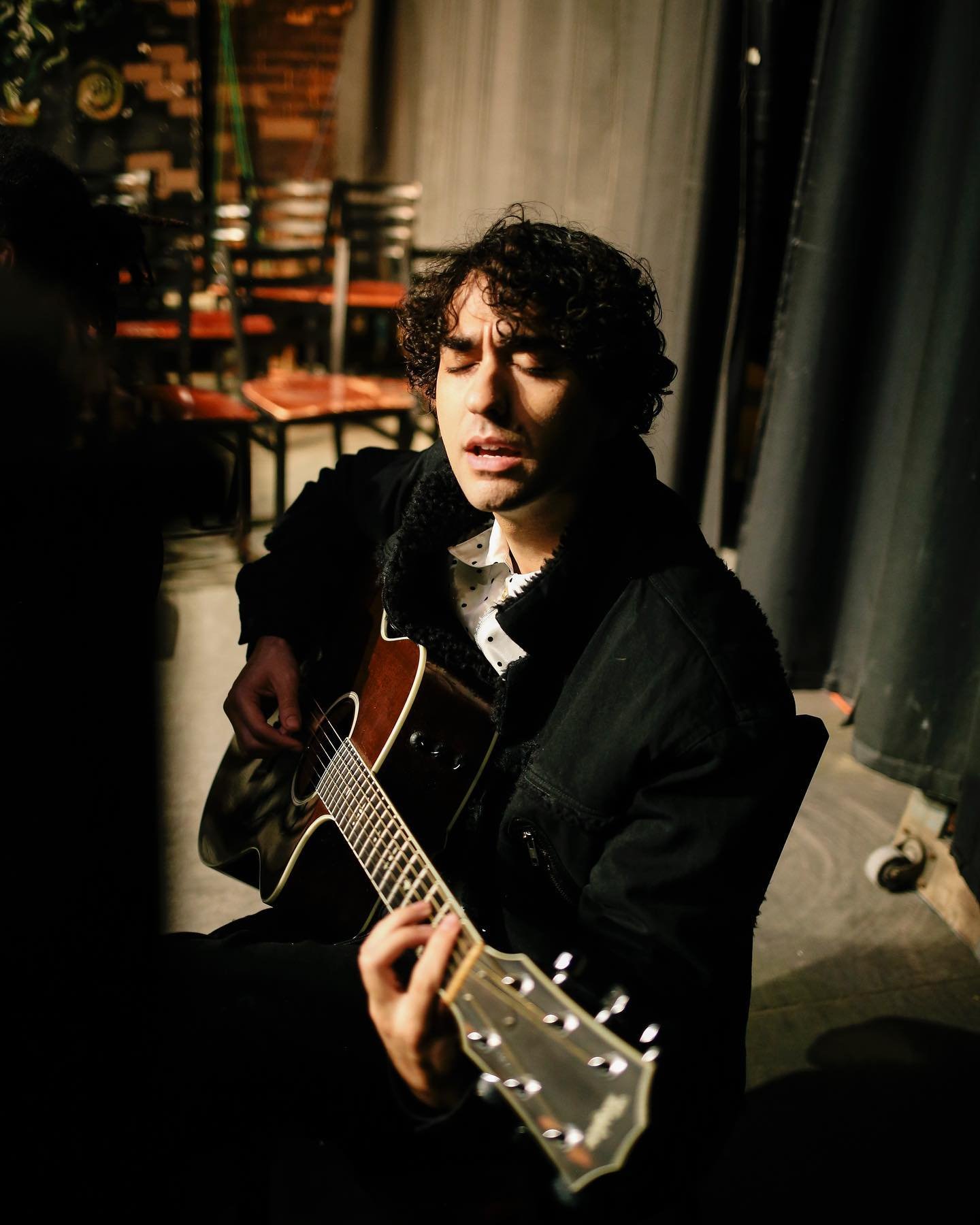 An adult man with curly hair sits and plays an acoustic guitar, wearing a black jacket and a patterned shirt in a dimly lit interior.