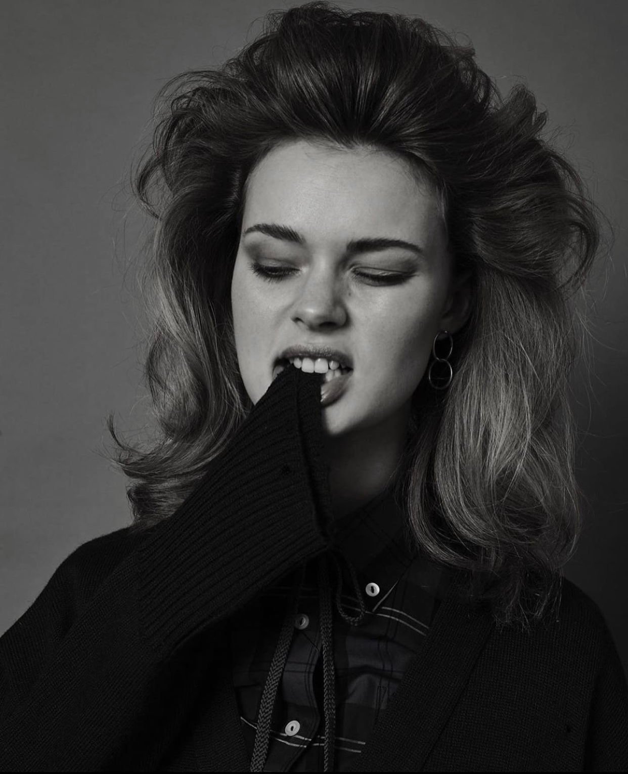 Monochrome portrait of a woman with voluminous hair biting her sleeve against a dark background