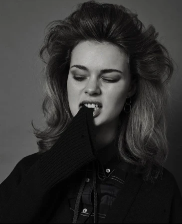 Monochrome portrait of a woman with voluminous hair biting her sleeve against a dark background