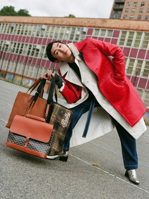 Stylish Asian man in red coat and silver boots holds multiple tote bags outdoors, posing in an urban environment with brick buildings.