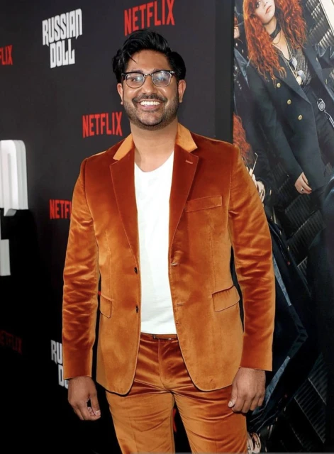 A man in a rust-colored velvet suit poses on a red carpet in front of a promotional poster for the Netflix series Russian Doll.