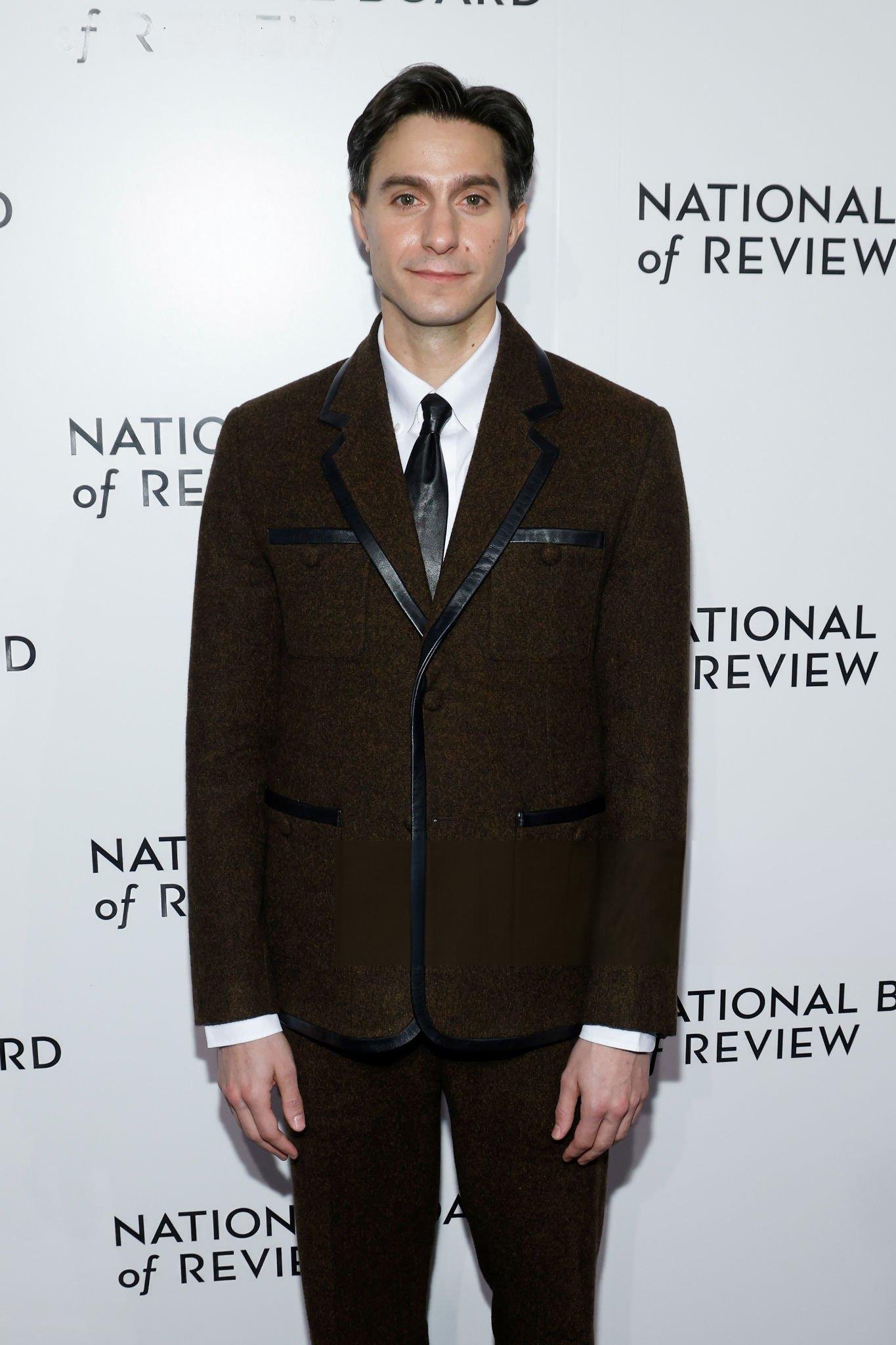 Man in a dark brown suit jacket with black piping standing against a white branded step and repeat backdrop.