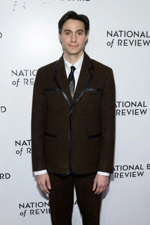 Man in a dark brown suit jacket with black piping standing against a white branded step and repeat backdrop.