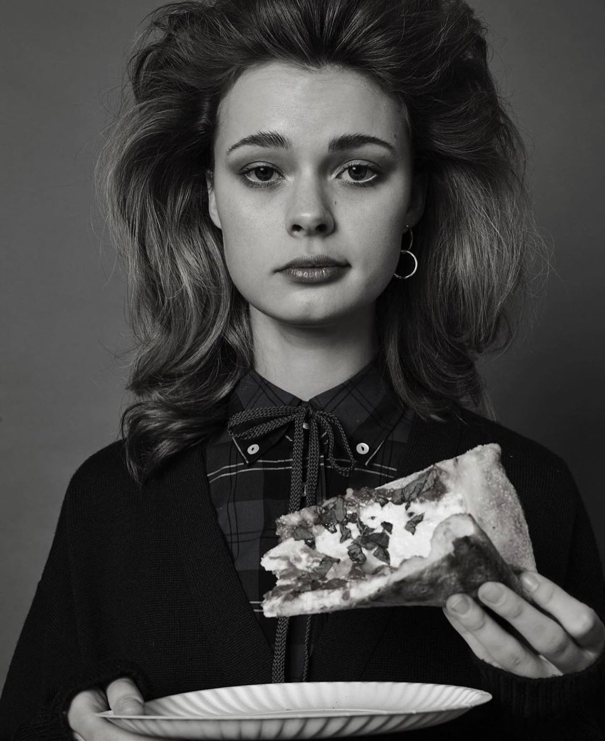 Monochrome portrait of a young woman holding a plate and pizza slice, wearing a plaid shirt and cardigan, against a grey backdrop.