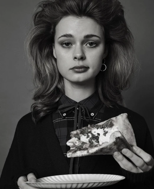 Monochrome portrait of a young woman holding a plate and pizza slice, wearing a plaid shirt and cardigan, against a grey backdrop.