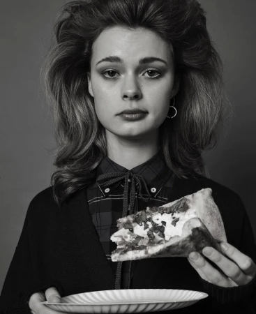 Monochrome portrait of a young woman holding a plate and pizza slice, wearing a plaid shirt and cardigan, against a grey backdrop.