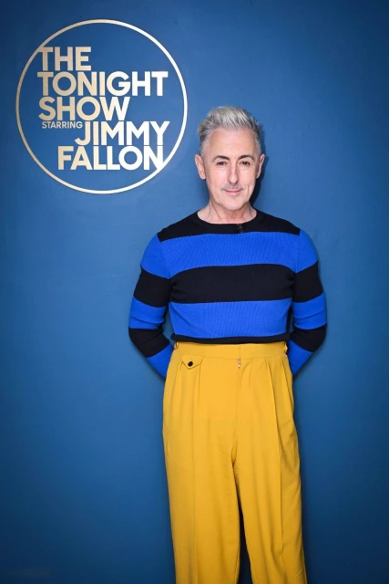Portrait of Alan Cumming in striped top and mustard pants, standing in front of 'The Tonight Show Starring Jimmy Fallon' logo.