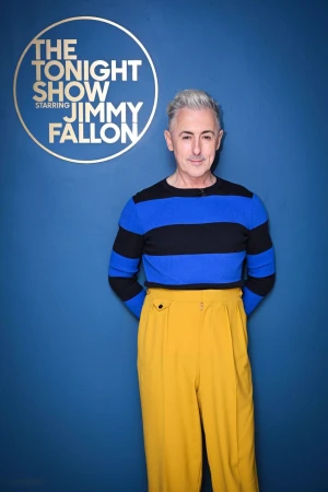 Portrait of Alan Cumming in striped top and mustard pants, standing in front of 'The Tonight Show Starring Jimmy Fallon' logo.