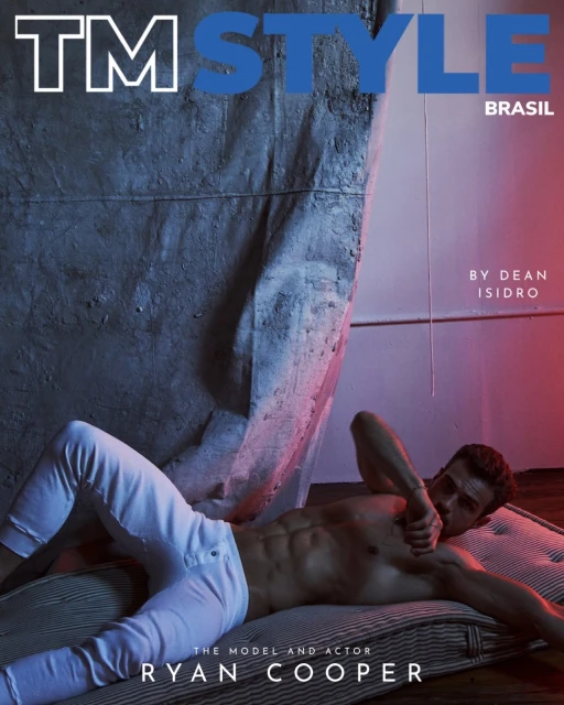 Shirtless man with abs lying on a bed wearing white pants, posing for TM Style Brasil magazine cover, grey concrete background.