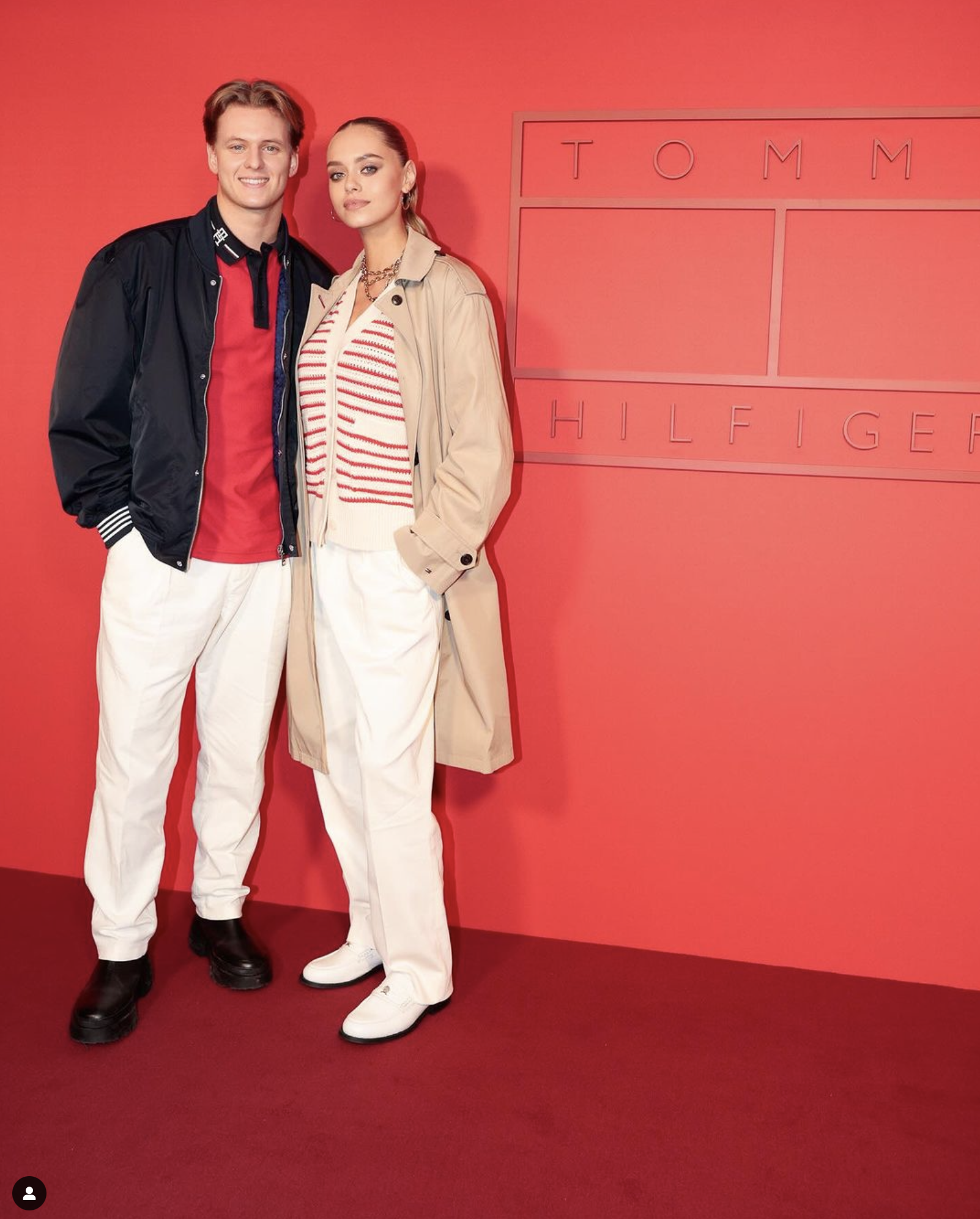 A fashionable couple posing in front of a Tommy Hilfiger sign, showcasing modern style and design against a red backdrop.