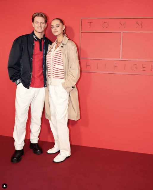 A fashionable couple posing in front of a Tommy Hilfiger sign, showcasing modern style and design against a red backdrop.