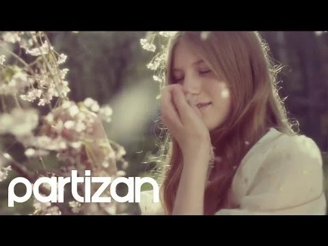 Young woman with long hair stands in a floral outdoor setting, wearing a white top and looking down in a close-up shot.