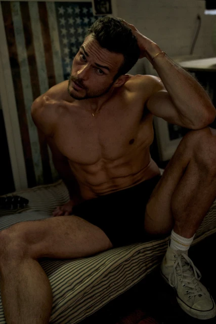 Male model poses shirtless with an American flag visible in the background. He sits on a striped surface with sneakers.