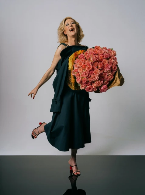 Smiling woman in dark dress and heels, balances on one leg holding a large bouquet of roses in a studio shot against grey.