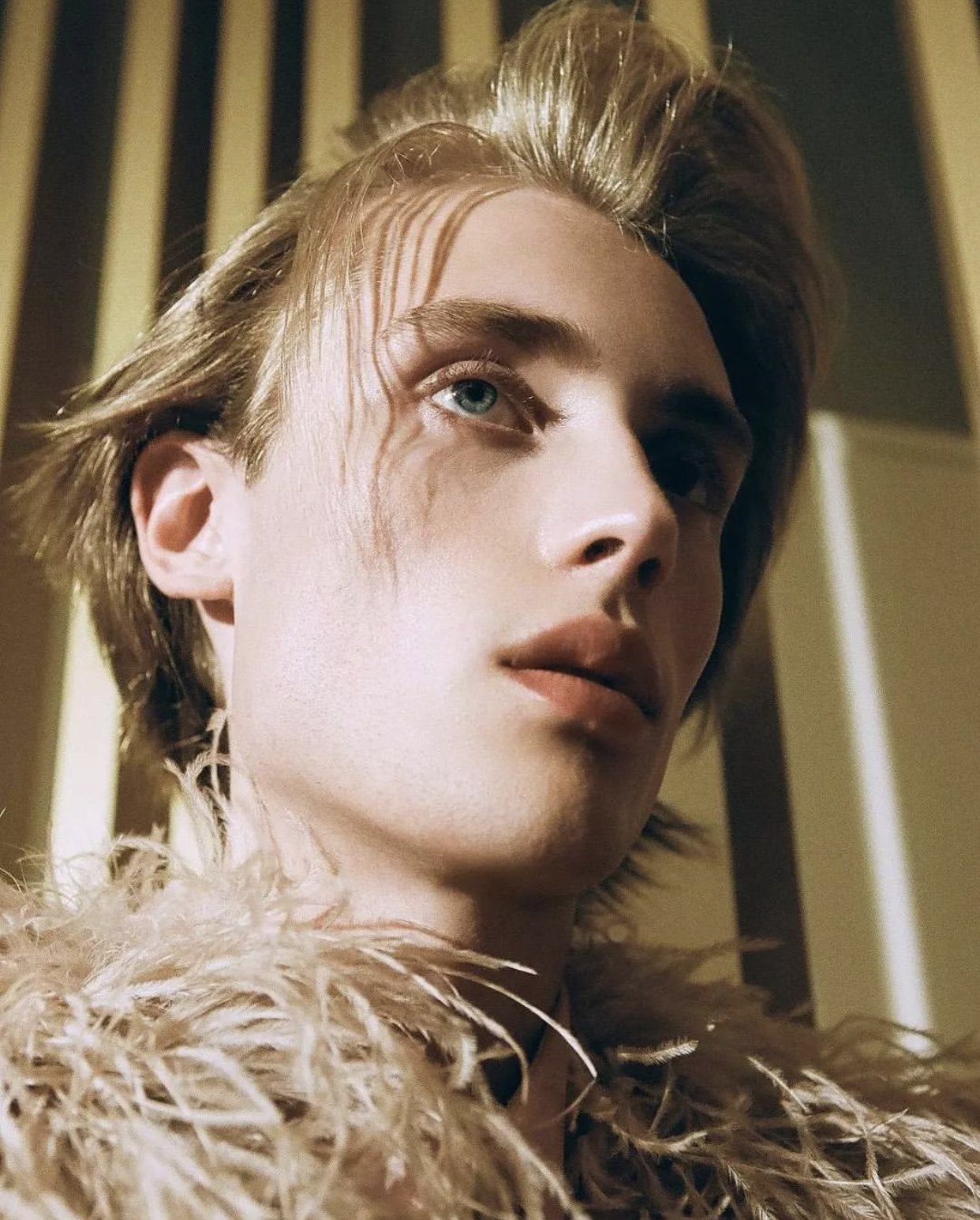 Close-up portrait of a young adult male with fair skin and blonde hair, wearing a feathered garment, against a background of striped lighting.