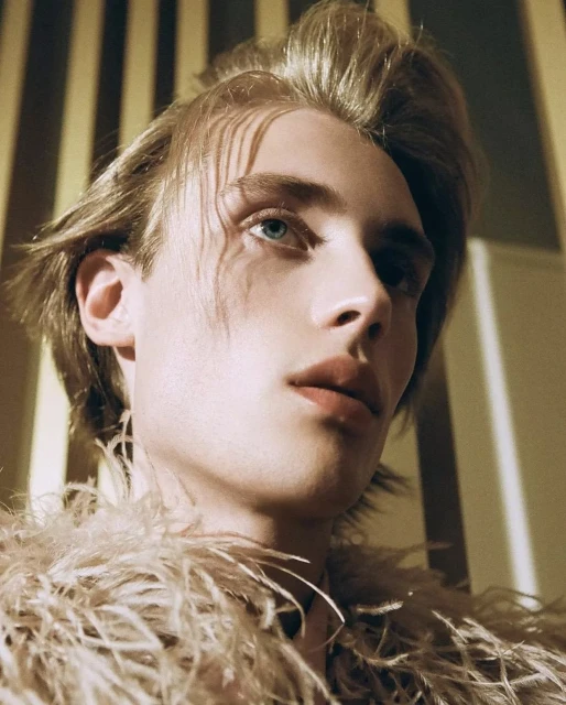 Close-up portrait of a young adult male with fair skin and blonde hair, wearing a feathered garment, against a background of striped lighting.