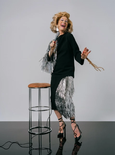 Adult female entertainer wearing a black dress with fringe, singing into a microphone, standing next to a stool in a studio.