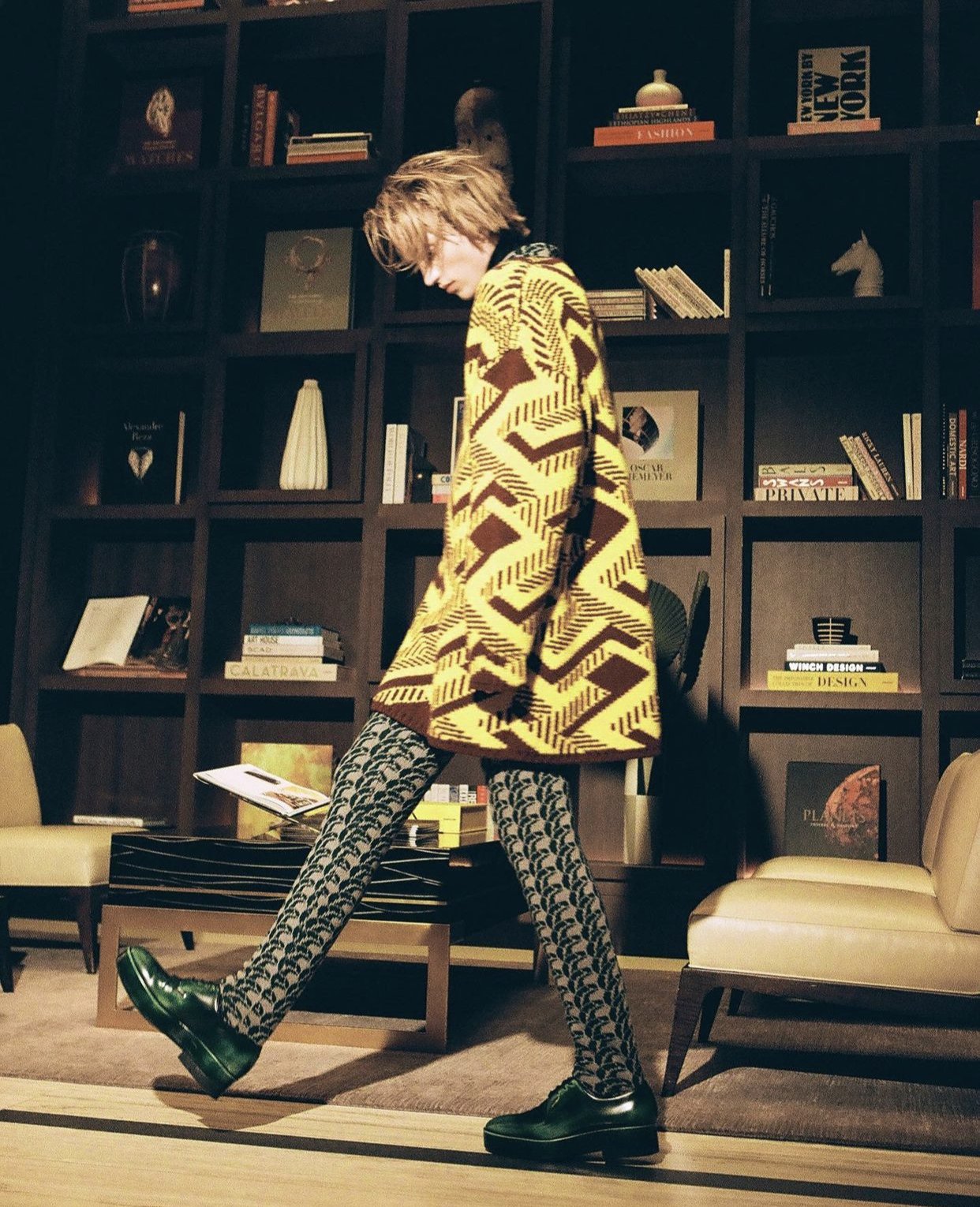 Fashionable adult in patterned cardigan and stockings walking in an interior setting with bookshelves, chairs, and decorative objects.