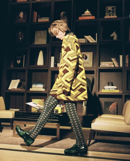 Fashionable adult in patterned cardigan and stockings walking in an interior setting with bookshelves, chairs, and decorative objects.