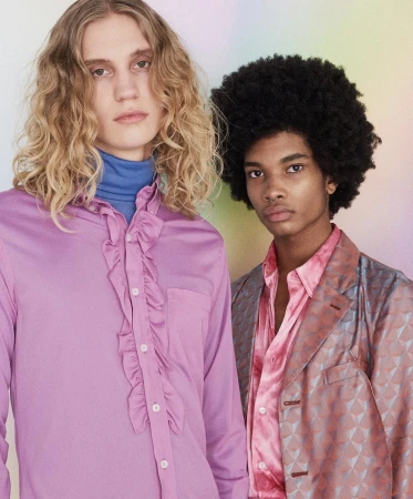 Two young men in a studio setting. One with blond wavy hair wears a lilac ruffled shirt. The other with afro hair, wears a pink patterned blazer.
