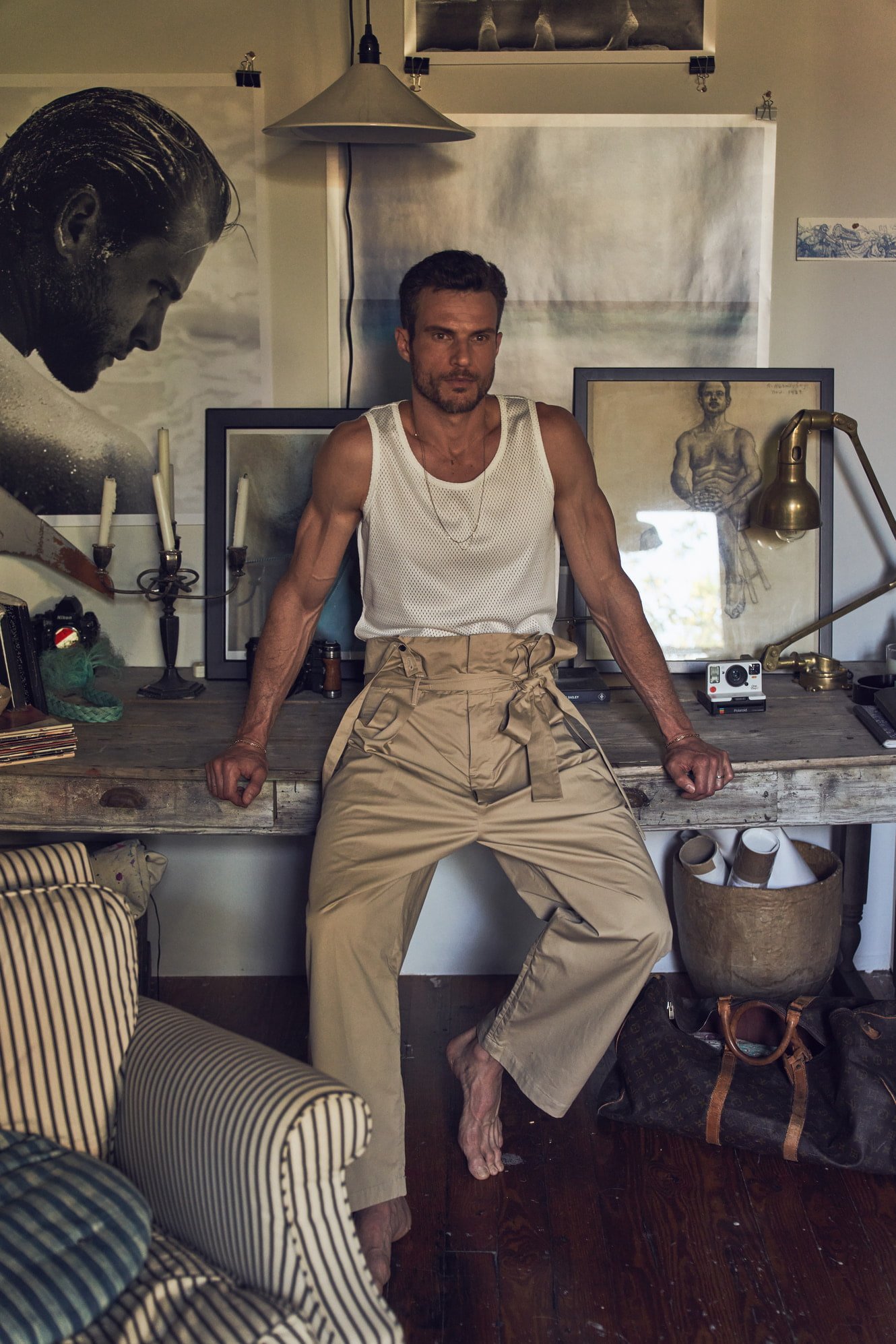 Adult man sitting and leaning on a rustic wooden desk in a bright room with artwork; wearing a white tank top and tan wide-legged pants.