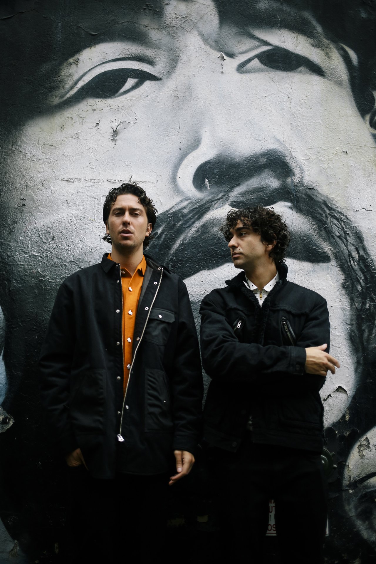 Two adult men stand in front of a graffiti mural featuring a large face on a weathered wall, captured in a medium shot.