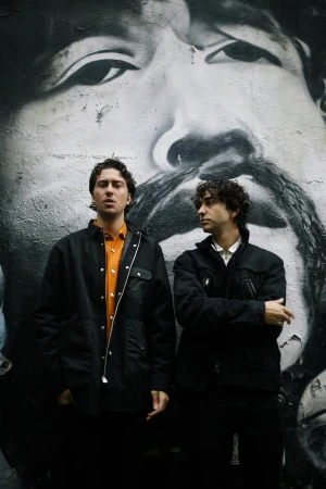 Two adult men stand in front of a graffiti mural featuring a large face on a weathered wall, captured in a medium shot.