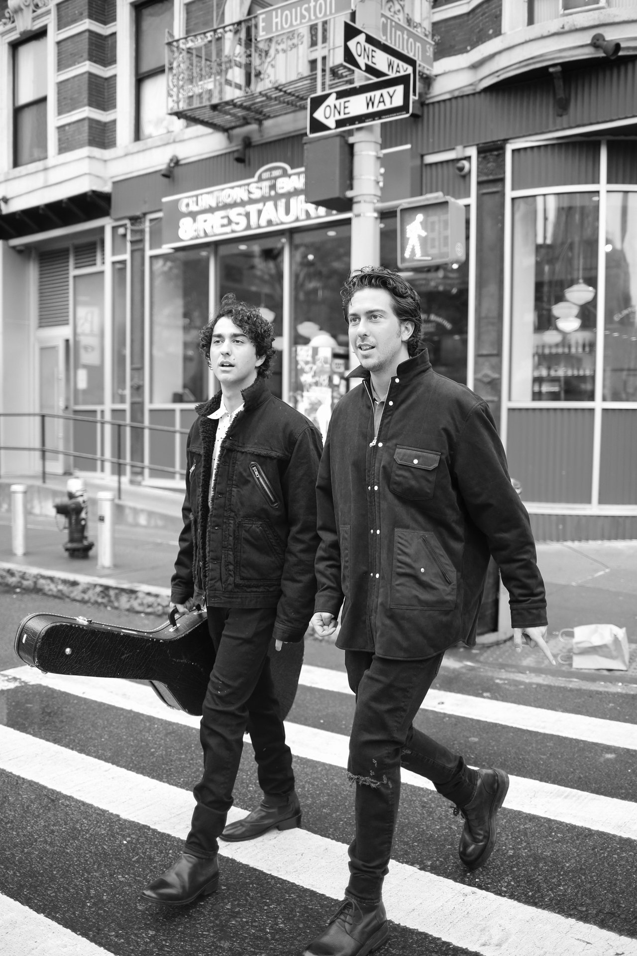 Two men in black jackets crossing the street in New York City, one carrying a guitar case, captured in black and white.