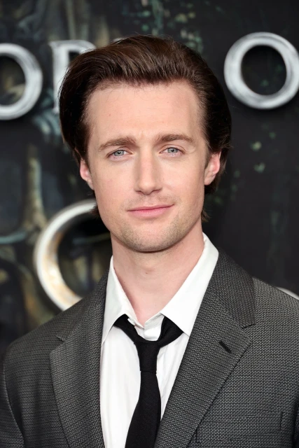 Close-up portrait of a dark-haired man in a grey suit jacket and black tie, standing against a blurred, dark background.