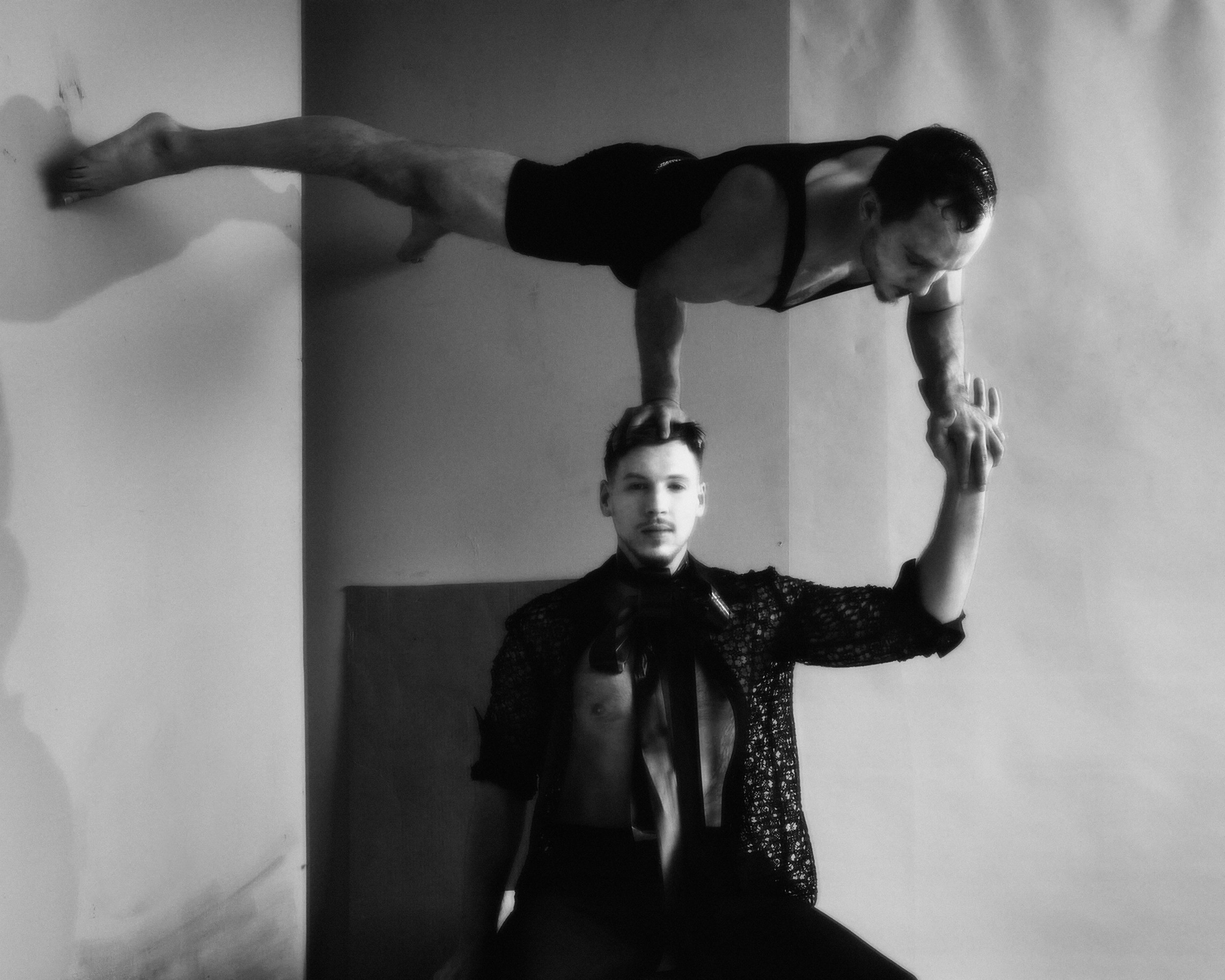 Two male acrobats in a black and white photo. One man balances on top of the other, striking a pose against a neutral background.