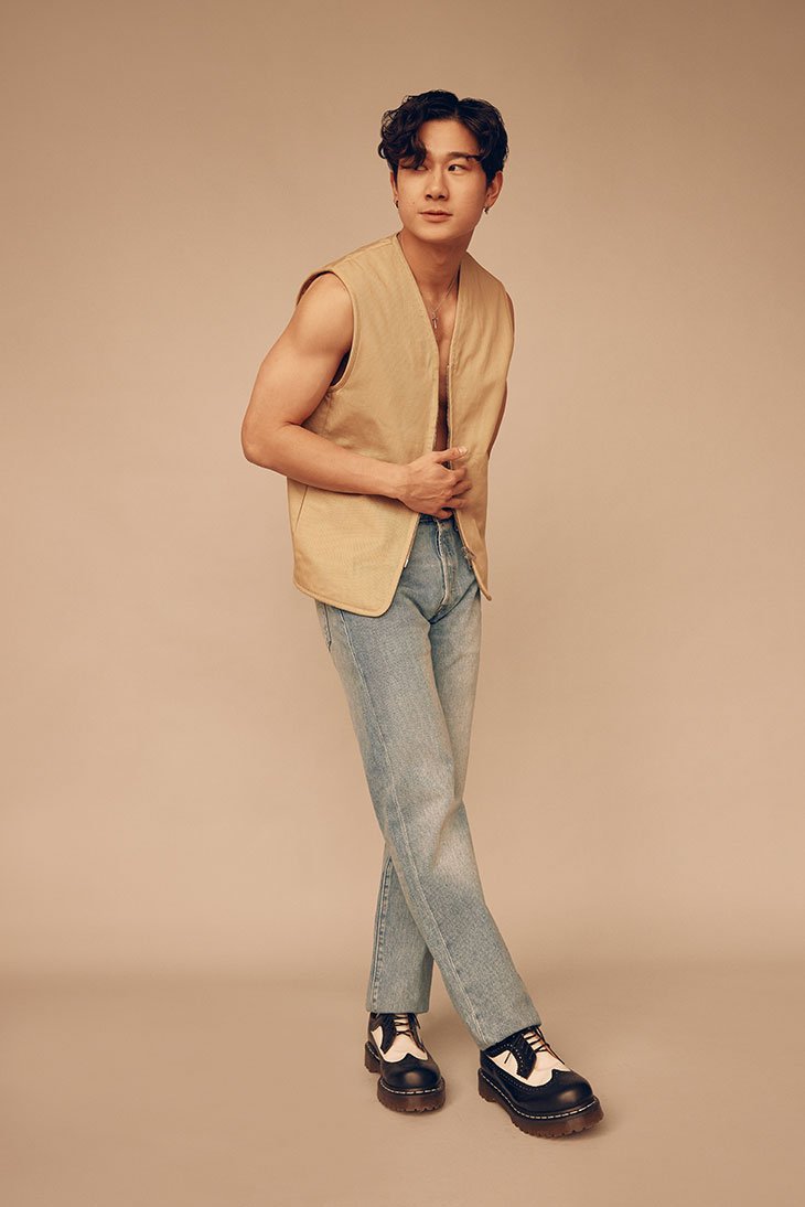 Portrait of an Asian man wearing a vest and jeans in a studio. He's standing with his arms crossed and looking off to the side.