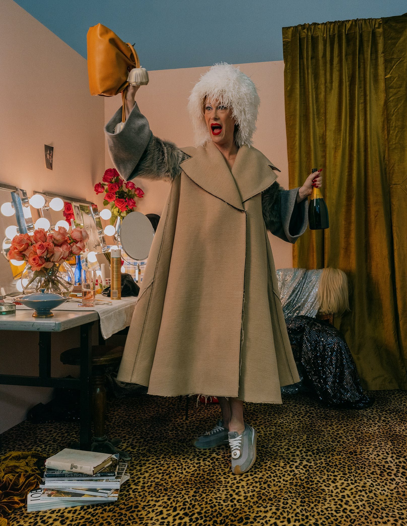 Adult female celebrates in a dressing room with champagne and a quirky outfit featuring an oversized coat and fuzzy white wig.