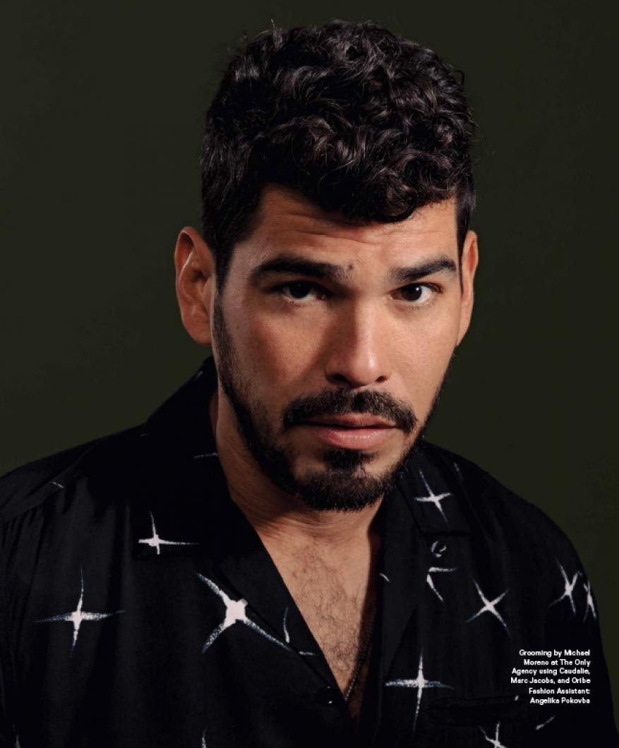Portrait of a man with dark curly hair and a beard, wearing a patterned shirt, against a dark green background. Studio shot.