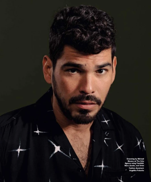Portrait of a man with dark curly hair and a beard, wearing a patterned shirt, against a dark green background. Studio shot.