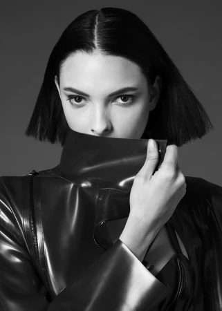 Close-up monochrome portrait of a woman with dark bobbed hair holding up the high collar of a glossy dark coat.