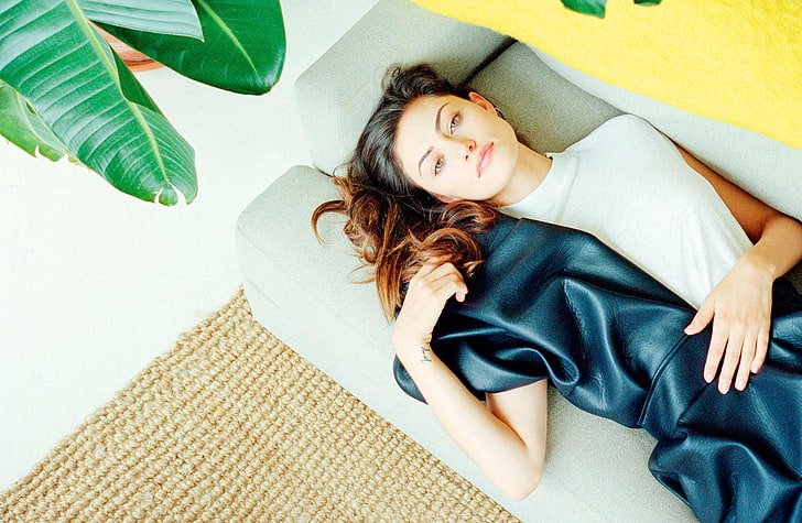 Brunette woman relaxes on a gray couch indoors with a black dress, light walls, woven rug, and houseplant adding natural, stylish vibes.