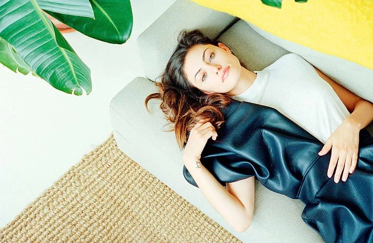 Brunette woman relaxes on a gray couch indoors with a black dress, light walls, woven rug, and houseplant adding natural, stylish vibes.