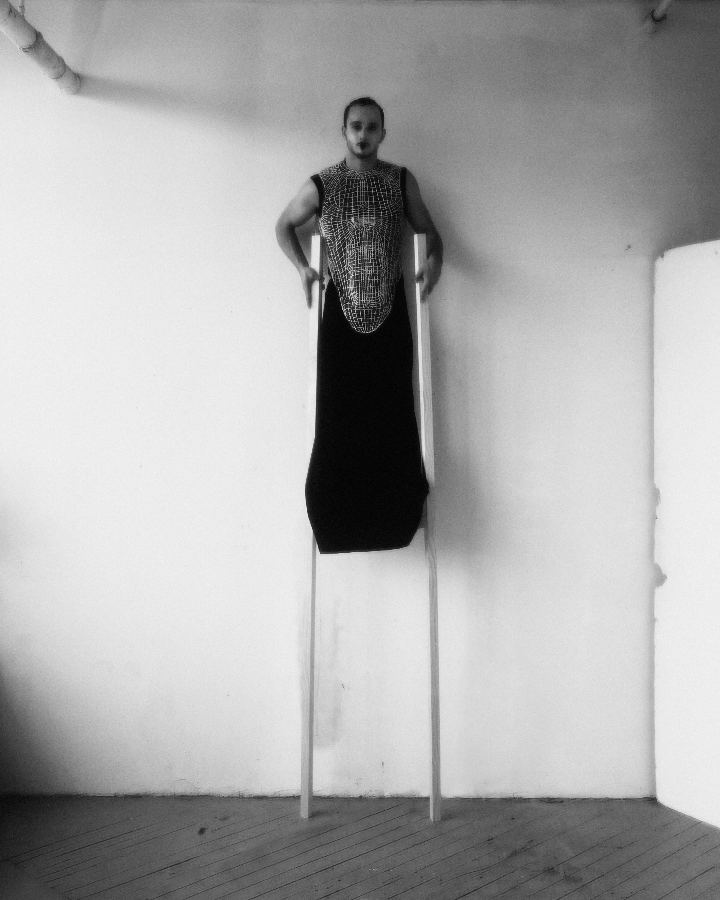 Black and white portrait of an adult male in a chainmail vest and dark dress, standing on stilts in a minimalist studio setting.