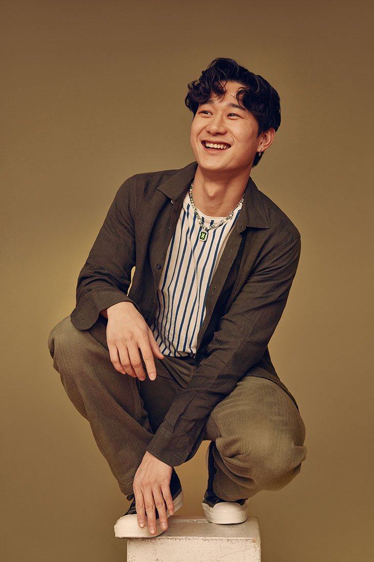Smiling Asian man in striped t-shirt and olive-green jacket squats on a white cube in a studio with olive-green back drop.