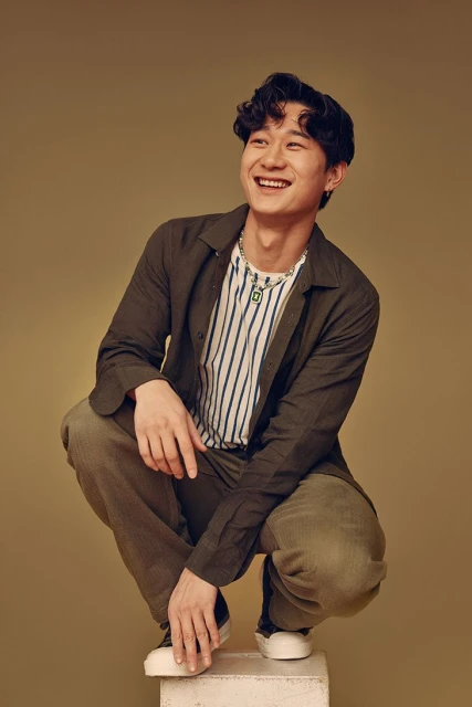 Smiling Asian man in striped t-shirt and olive-green jacket squats on a white cube in a studio with olive-green back drop.
