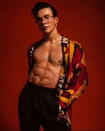 Portrait of a man in a patterned yellow, red, and black unbuttoned shirt and dark trousers against a warm red background.