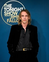 Jamie Campbell Bower in a black velvet suit jacket against The Tonight Show backdrop with hair styled by Michael Moreno