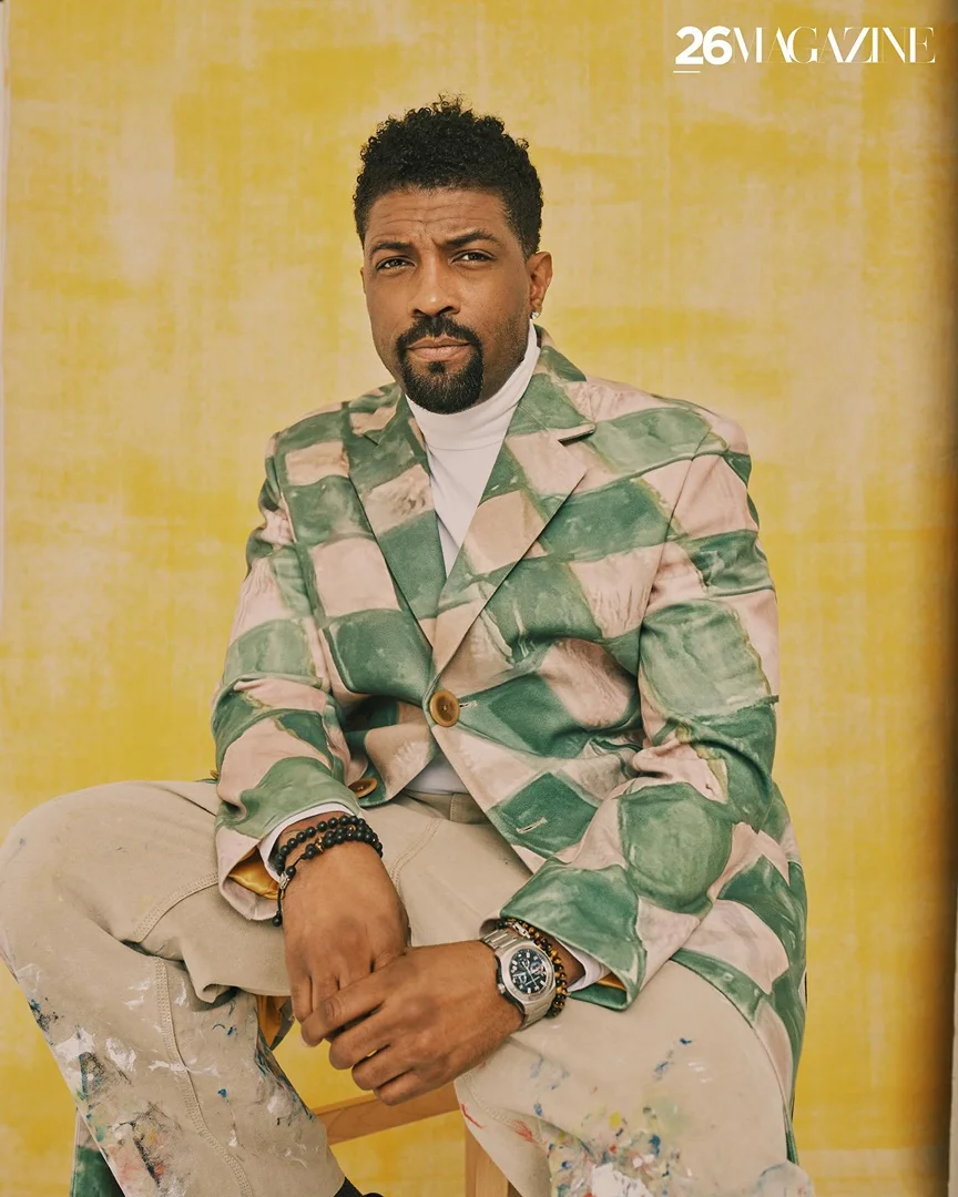 Portrait of a man seated, wearing a patterned jacket and paint-splattered pants, with a yellow textured background and '26 Magazine' logo.
